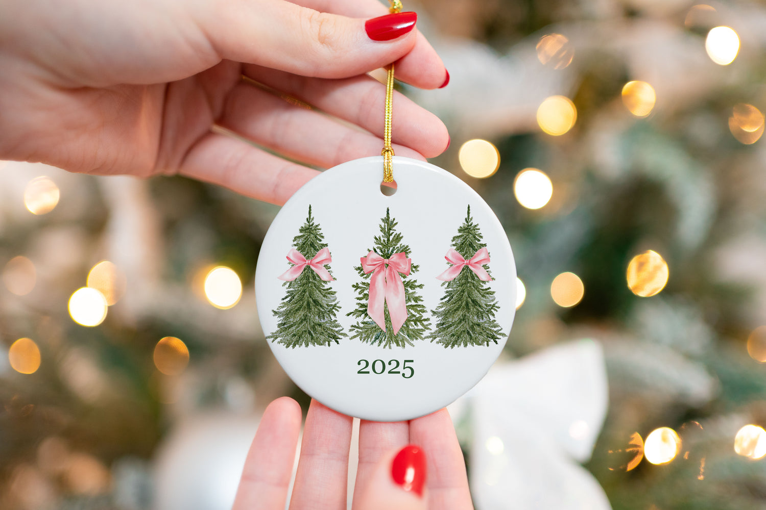 2025 Christmas ornament with three green watercolor trees, each tied with soft pink bows, professionally printed on a round ceramic base with gold string, held in front of a blurred Christmas tree background.
