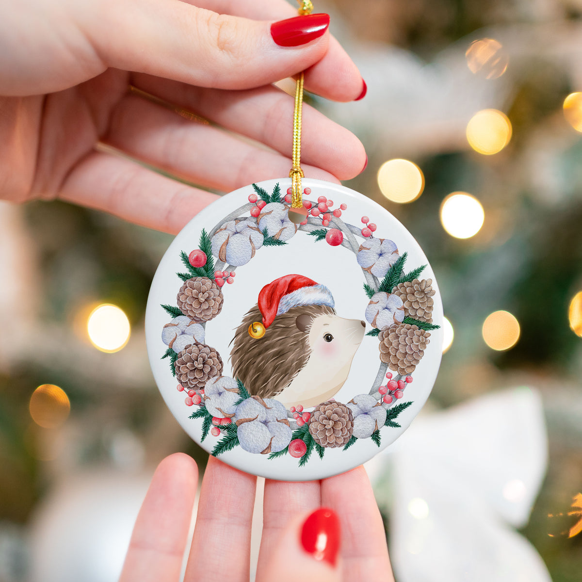 Close-up of a ceramic Christmas ornament featuring a hedgehog in a Santa hat surrounded by a holiday wreath with pinecones, berries, and cotton flowers. Ornament is held in front of a softly lit Christmas tree.