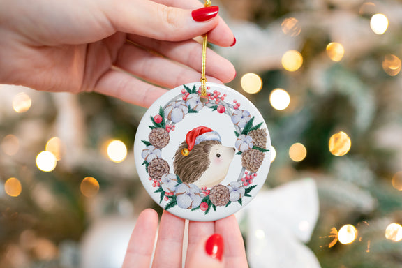 Close-up of a ceramic Christmas ornament featuring a hedgehog in a Santa hat surrounded by a holiday wreath with pinecones, berries, and cotton flowers. Ornament is held in front of a softly lit Christmas tree.