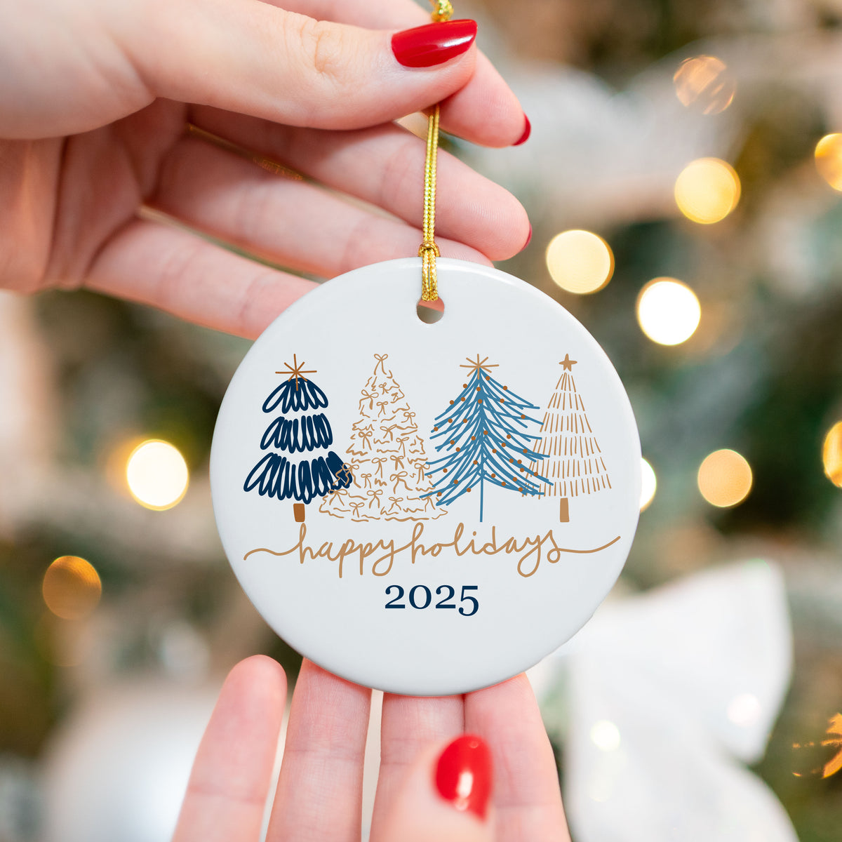 Hand holding a round white ceramic ornament with navy and gold modern Christmas trees and "Happy Holidays 2025" text, in front of a blurred Christmas tree with lights