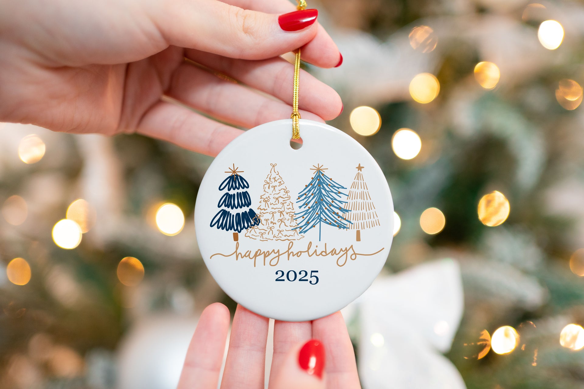 Hand holding a round white ceramic ornament with navy and gold modern Christmas trees and "Happy Holidays 2025" text, in front of a blurred Christmas tree with lights