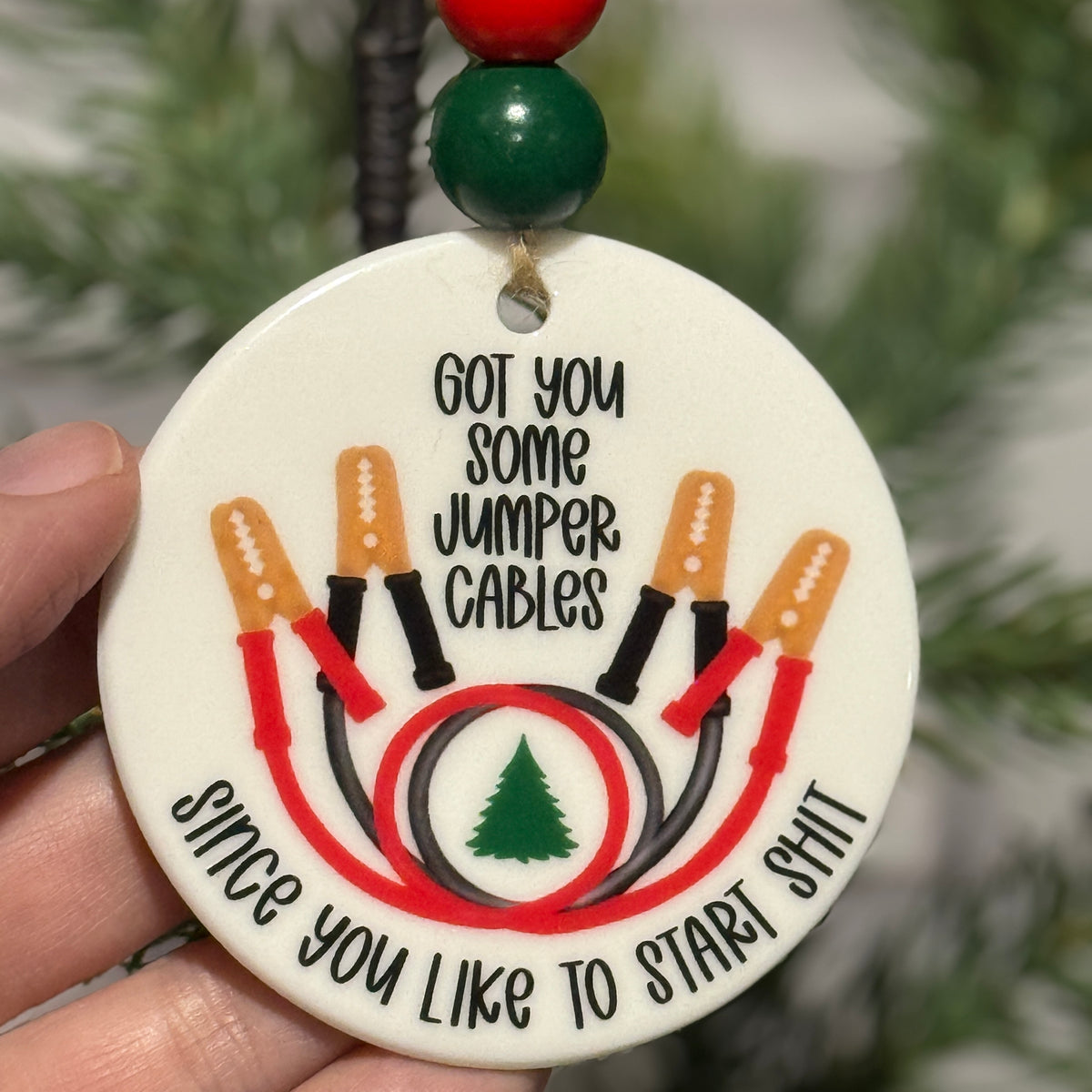Decorative ornament with jumper cables design held in front of a Christmas tree.