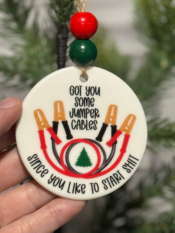 Decorative ornament with jumper cables design held in front of a Christmas tree.
