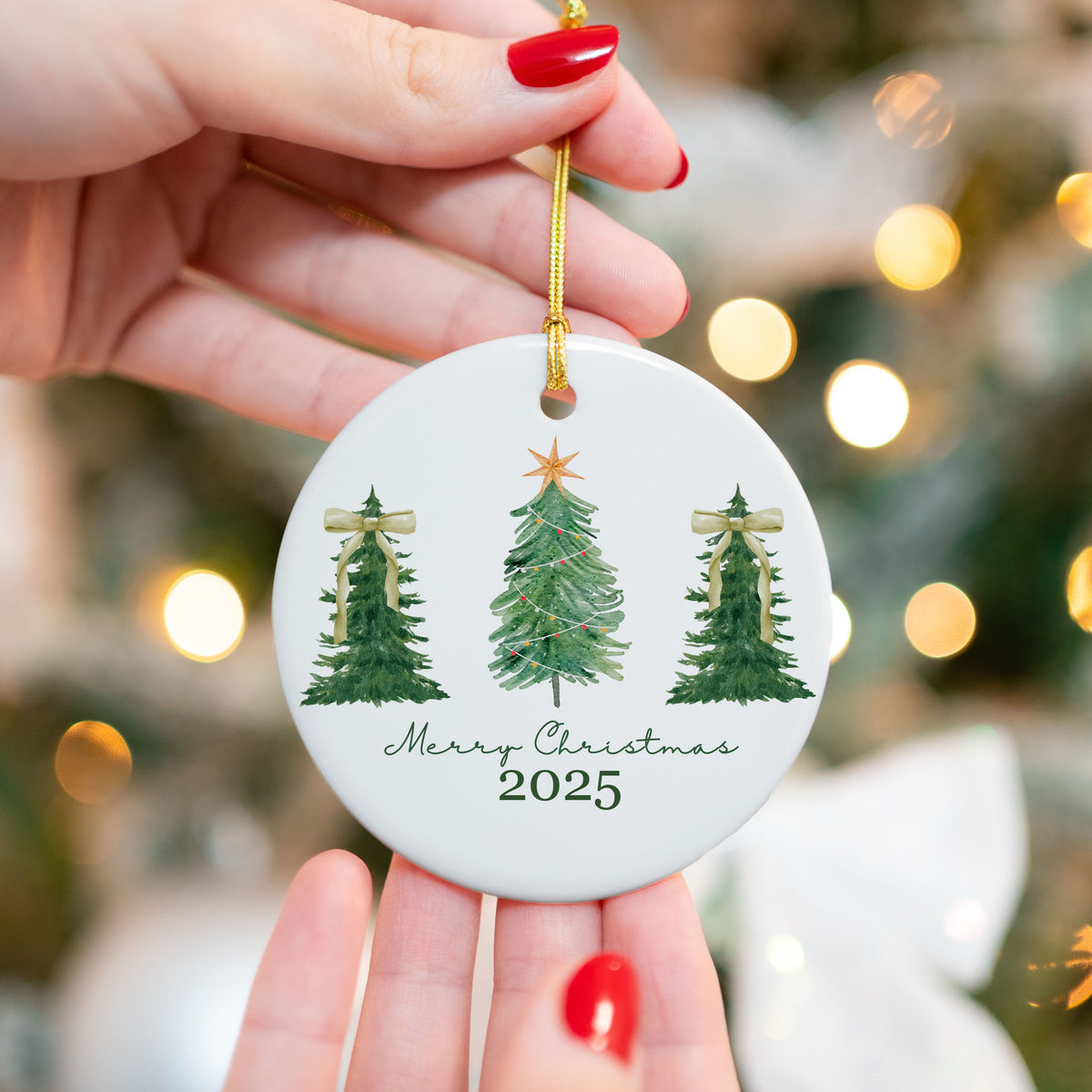 Ceramic Christmas ornament featuring three green Christmas trees with gold accents and the text "Merry Christmas 2025," held in front of a lit Christmas tree background.