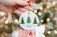 Ceramic Christmas ornament featuring three green Christmas trees with gold accents and the text "Merry Christmas 2025," held in front of a lit Christmas tree background.