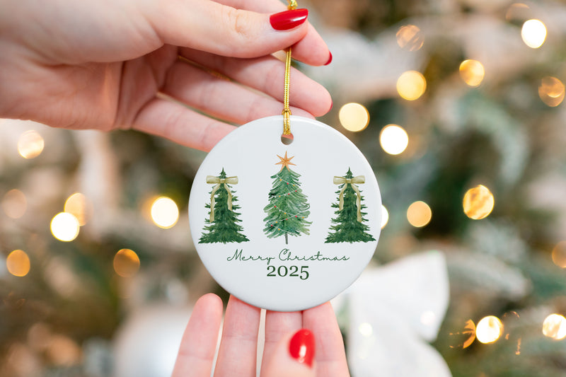 Ceramic Christmas ornament featuring three green Christmas trees with gold accents and the text "Merry Christmas 2025," held in front of a lit Christmas tree background.