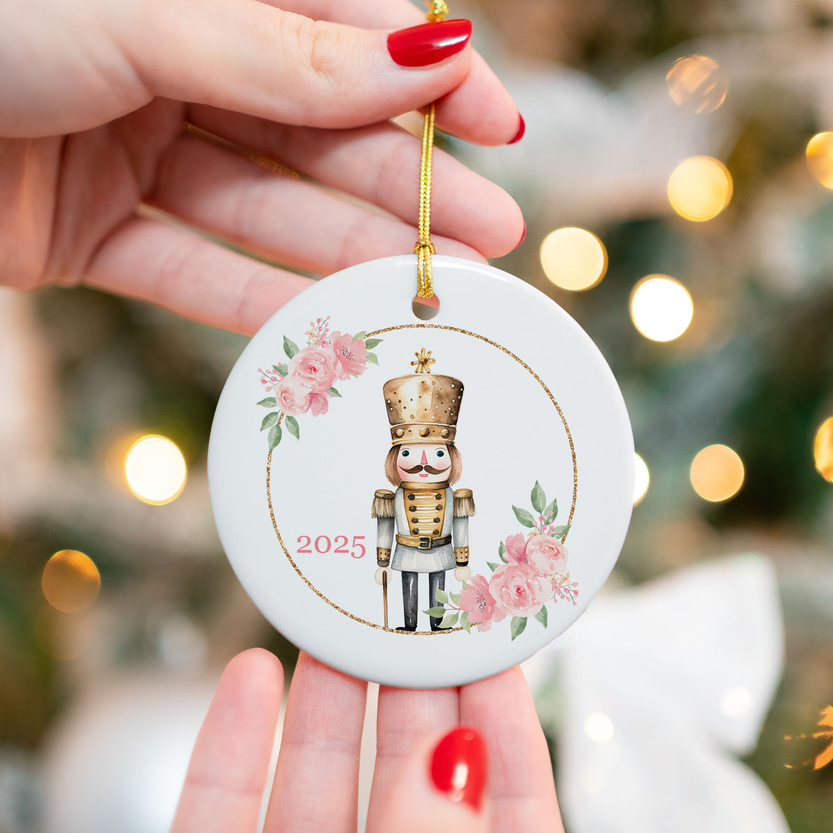 Close-up of a ceramic Christmas ornament featuring a gold nutcracker framed by pink roses and greenery, with “2025” printed on the front. Ornament is held in front of a Christmas tree with bokeh lights.