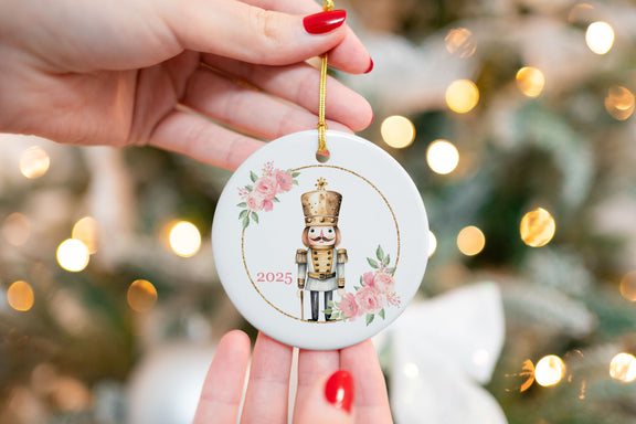 Close-up of a ceramic Christmas ornament featuring a gold nutcracker framed by pink roses and greenery, with “2025” printed on the front. Ornament is held in front of a Christmas tree with bokeh lights.