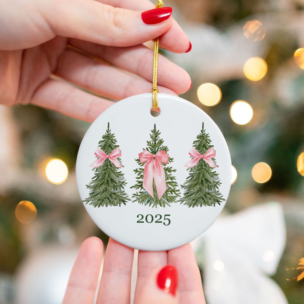 2025 Christmas ornament with three green watercolor trees, each tied with soft pink bows, professionally printed on a round ceramic base with gold string, held in front of a blurred Christmas tree background.